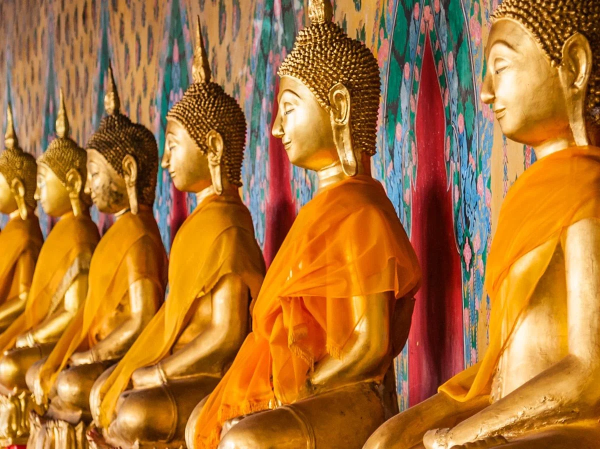 Line of golden Buddha statues wearing orange robes, seated against a colorful, patterned background.