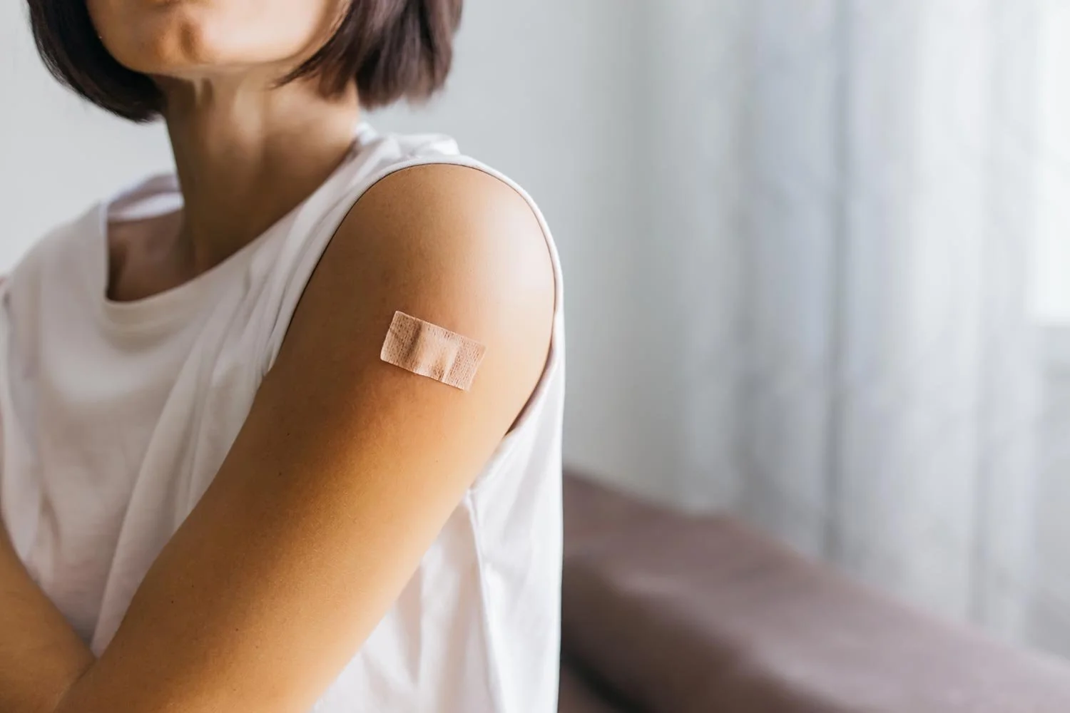 A person with short dark hair and wearing a white sleeveless shirt shows an arm with a bandage on the upper arm, indicating they have recently received a shot or vaccination, sitting indoors near a window with white curtains.