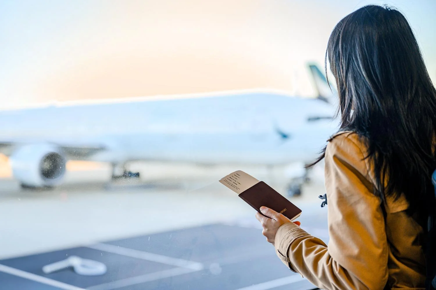 Woman waiting at airport gate looking at her boarding pass with airplane visible through the window in the background.