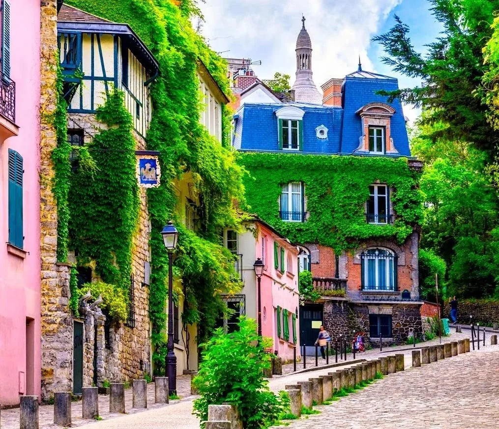 Colorful houses along a cobblestone street with green vines and trees, a lamp post, and a church steeple in the background.