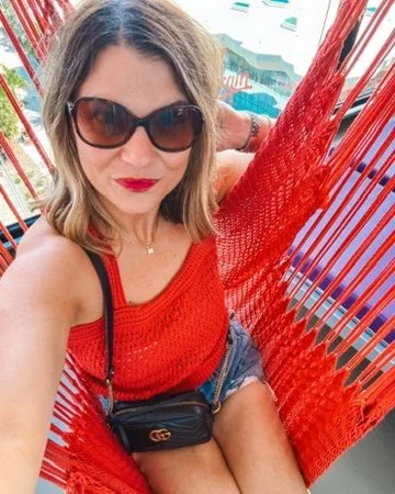 Woman with sunglasses wearing a red top and shorts, sitting in a red chair outdoors.