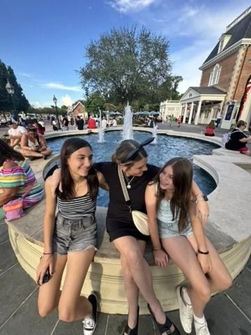 A woman and two young girls sitting on the edge of a circular fountain in a busy outdoor shopping area, smiling and enjoying the moment.
