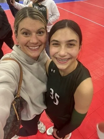 Two girls smiling and taking a selfie on a red indoor sports court. One girl has braces, and the other is wearing a gray hoodie. They appear happy and are close together.