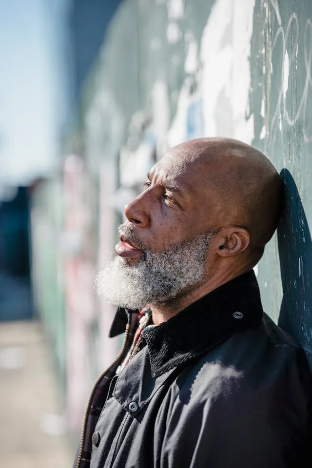 A man standing and leaning his back against a cement wall outdoors with side view of his face as he looks into the distance in thought