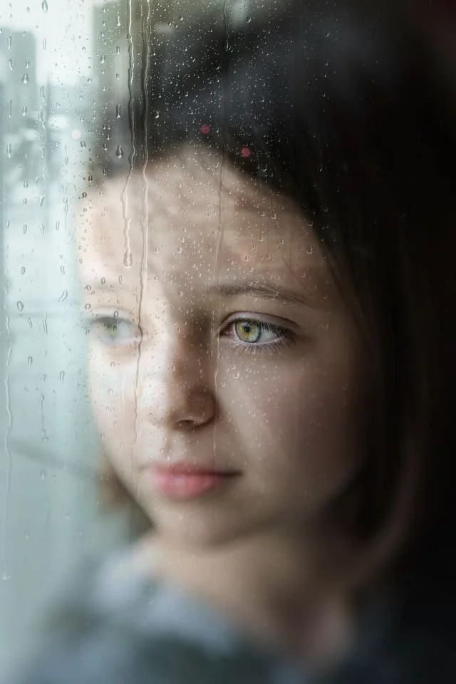 Young girl indoors sitting against and looking out rain covered window with thoughtful expression