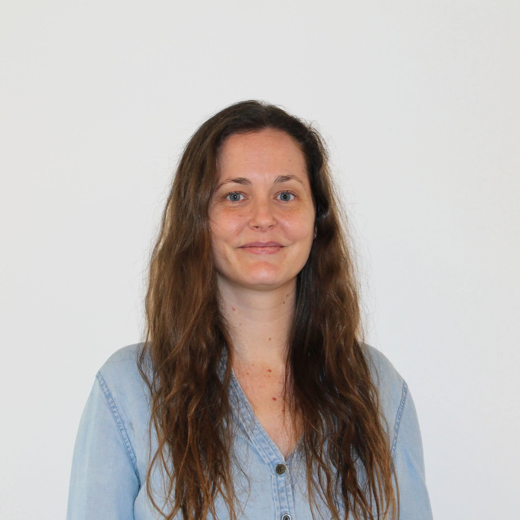 Portrait of a woman with long wavy brown hair, wearing a light blue denim shirt, standing against a plain white wall.