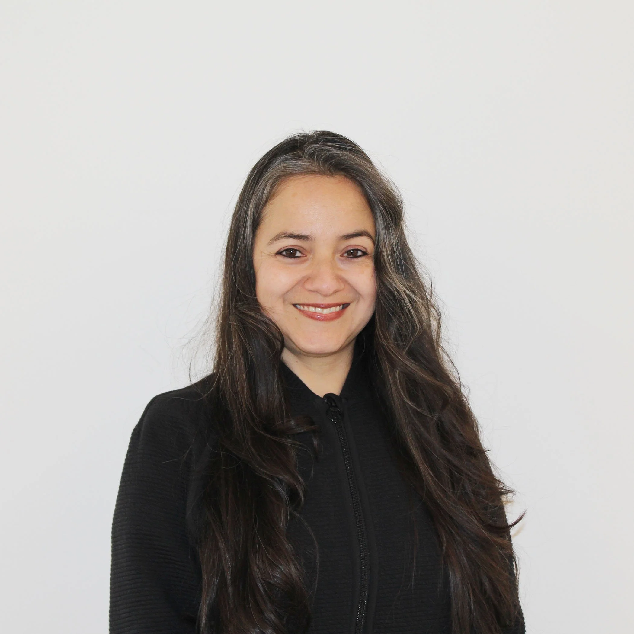 A woman with long dark hair, smiling, wearing a black top, standing against a plain off-white background.