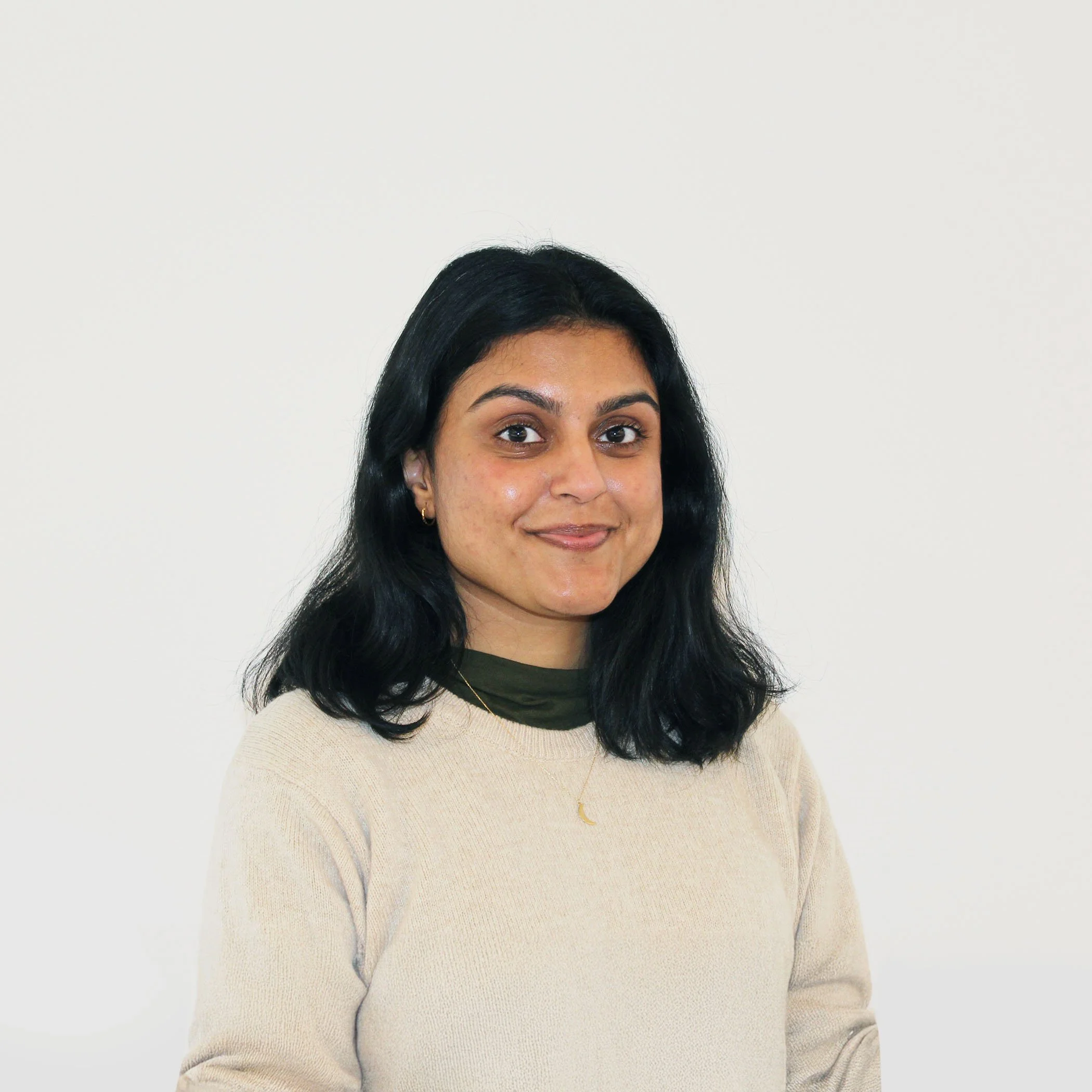 A young woman with shoulder-length black hair, dark eyebrows, and wearing a beige sweater, a dark green turtleneck underneath, small earrings, and a thin gold necklace with a crescent moon pendant, standing against a plain white wall.