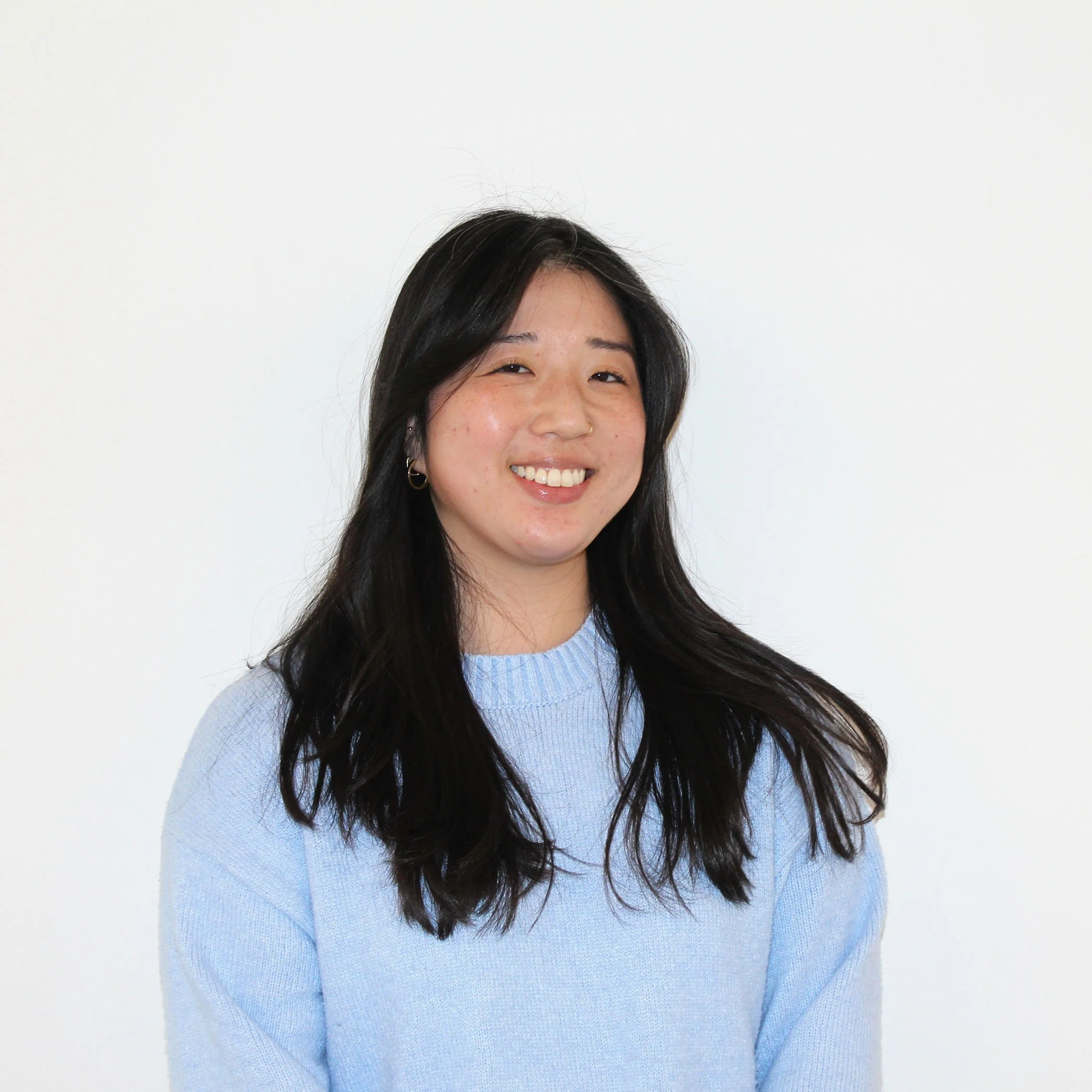 A young woman with long dark hair smiling, wearing a light blue sweater, standing against a plain white background.