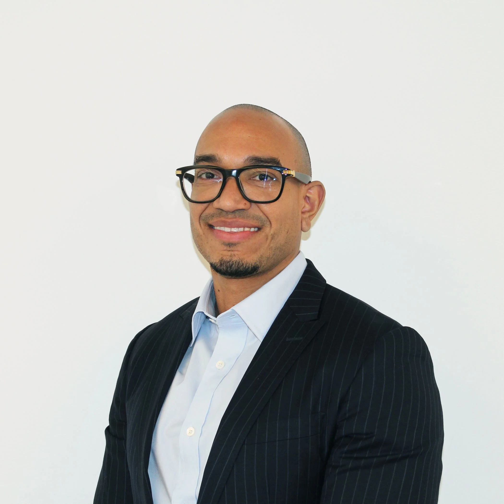 Portrait of a bald man with glasses, wearing a dark pinstripe suit and a light blue shirt, smiling against a plain off-white background.