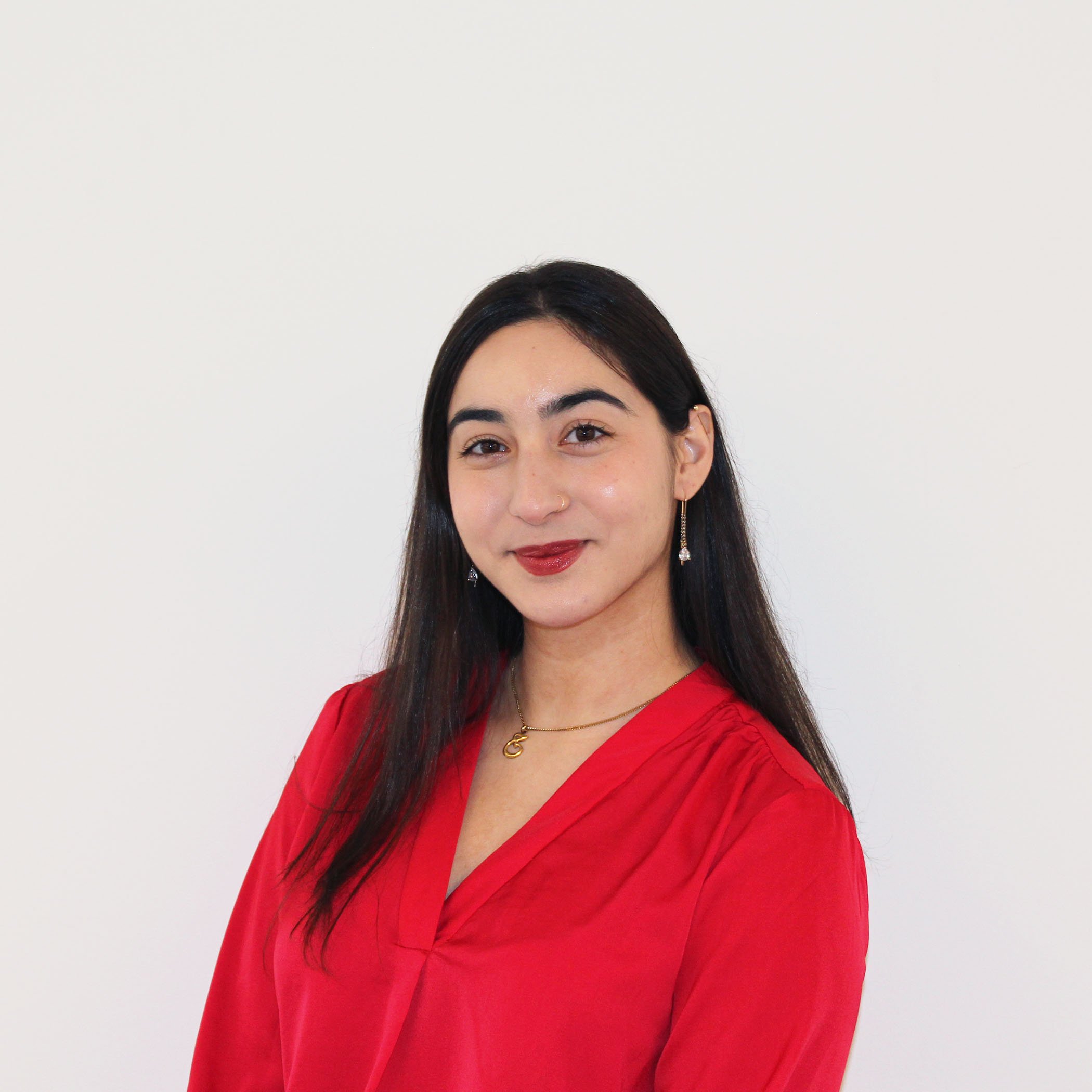 A woman with long dark hair wearing a red blouse, earrings, and lipstick, standing against a plain white wall.