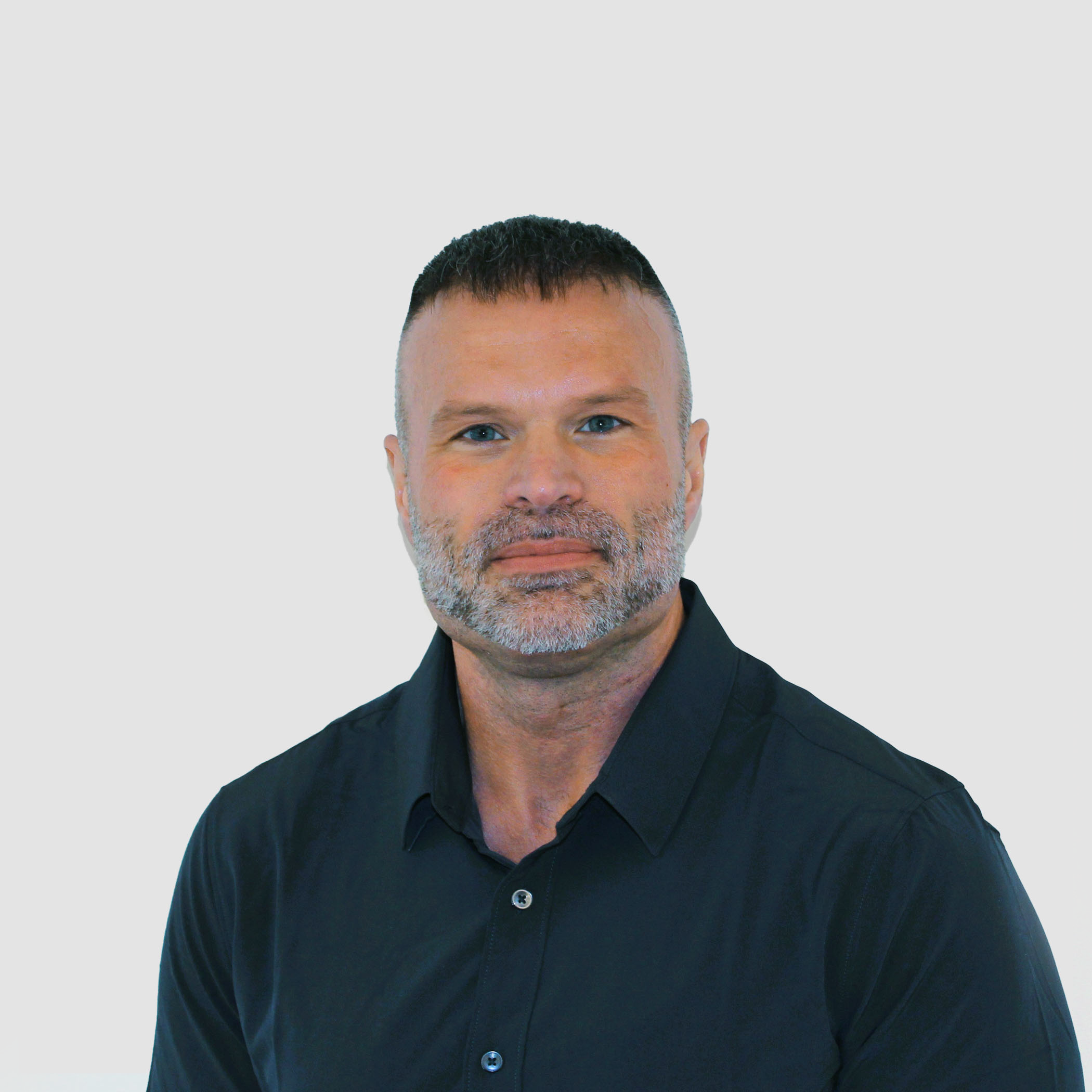 Man with short dark hair and beard, wearing a black button-up shirt, standing against a plain white wall.