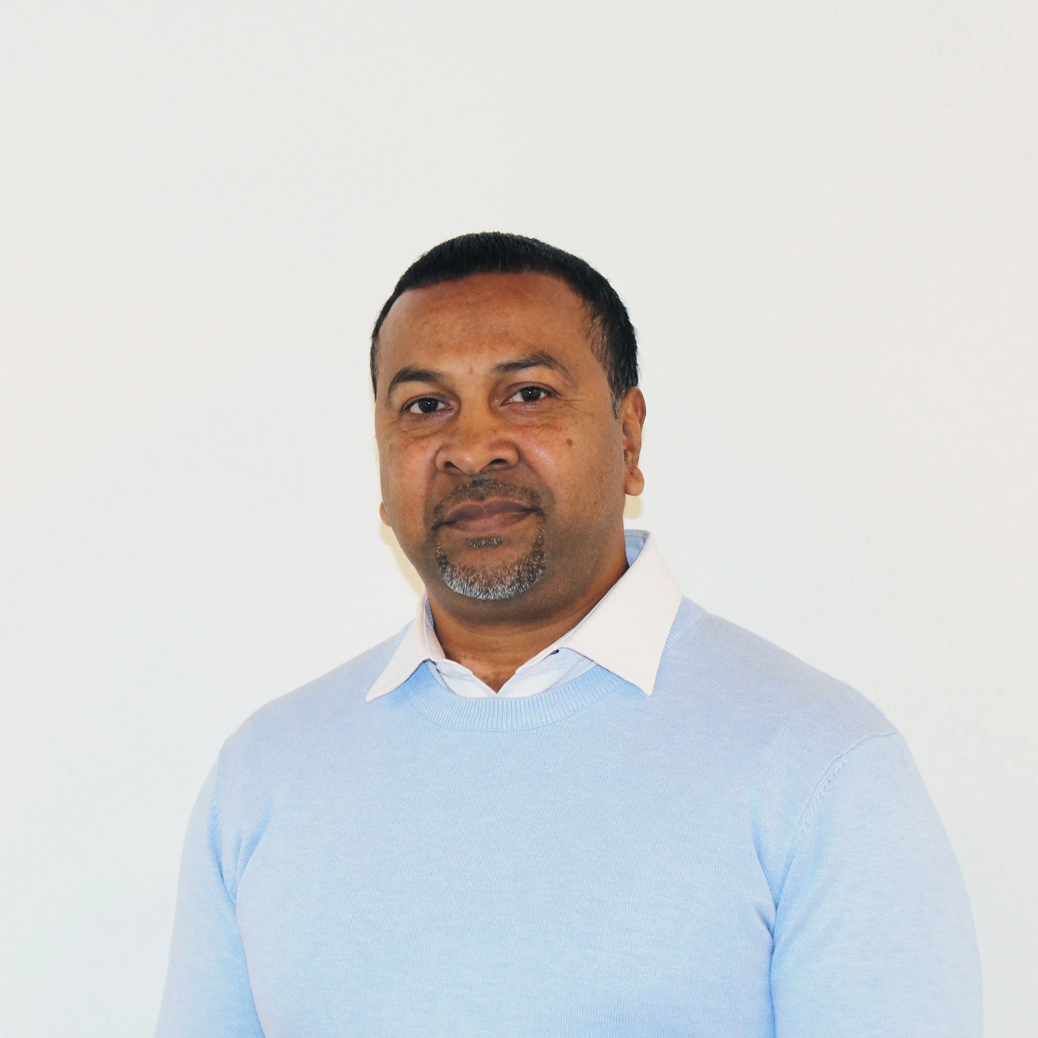 A man with short black hair, a goatee, and brown skin wearing a white collared shirt and a light blue sweater, standing against a plain white wall.