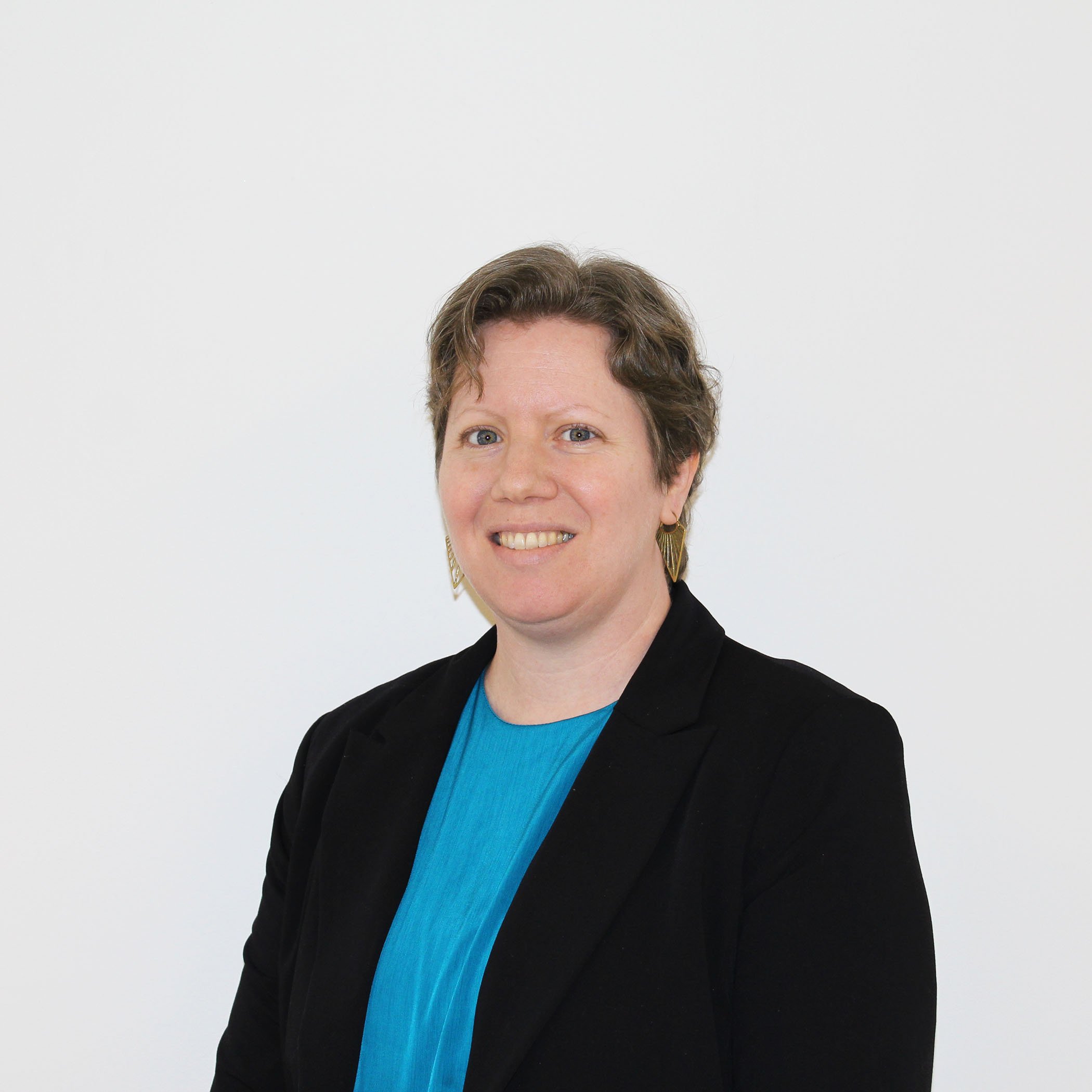 A woman with short brown hair smiling, wearing a black blazer over a bright blue top, standing against a plain white background.