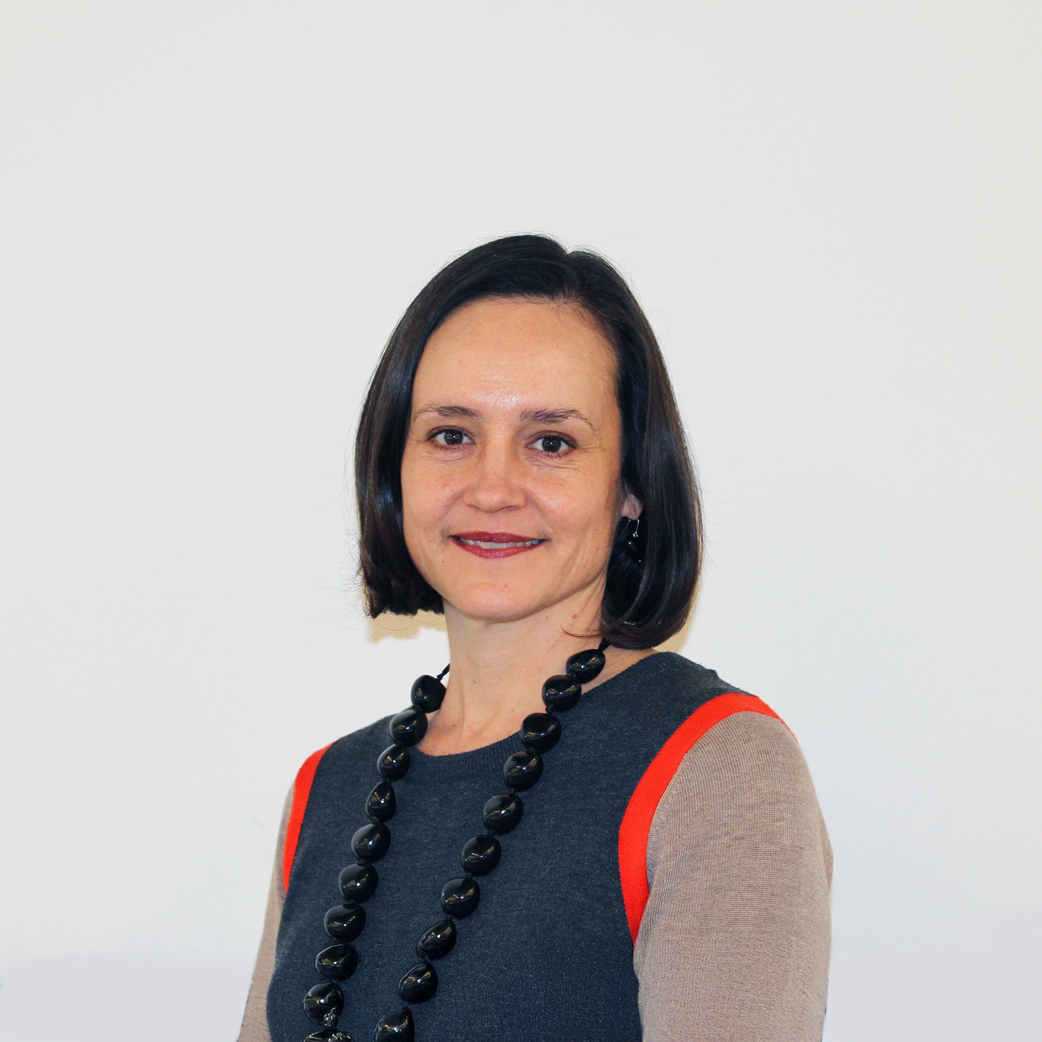 A woman with shoulder-length dark hair, wearing a gray and beige top with red accents, a black beaded necklace, and earrings, standing against a plain white background.