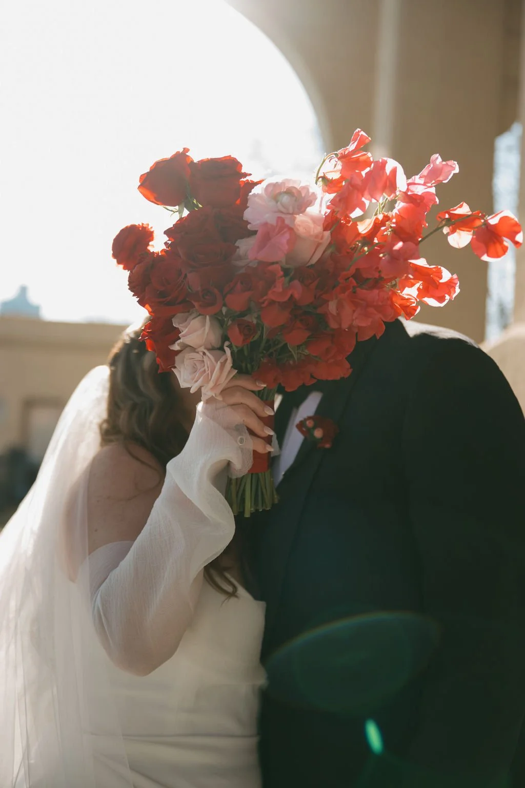 A couple kissing behind the brides red and pink bouquet