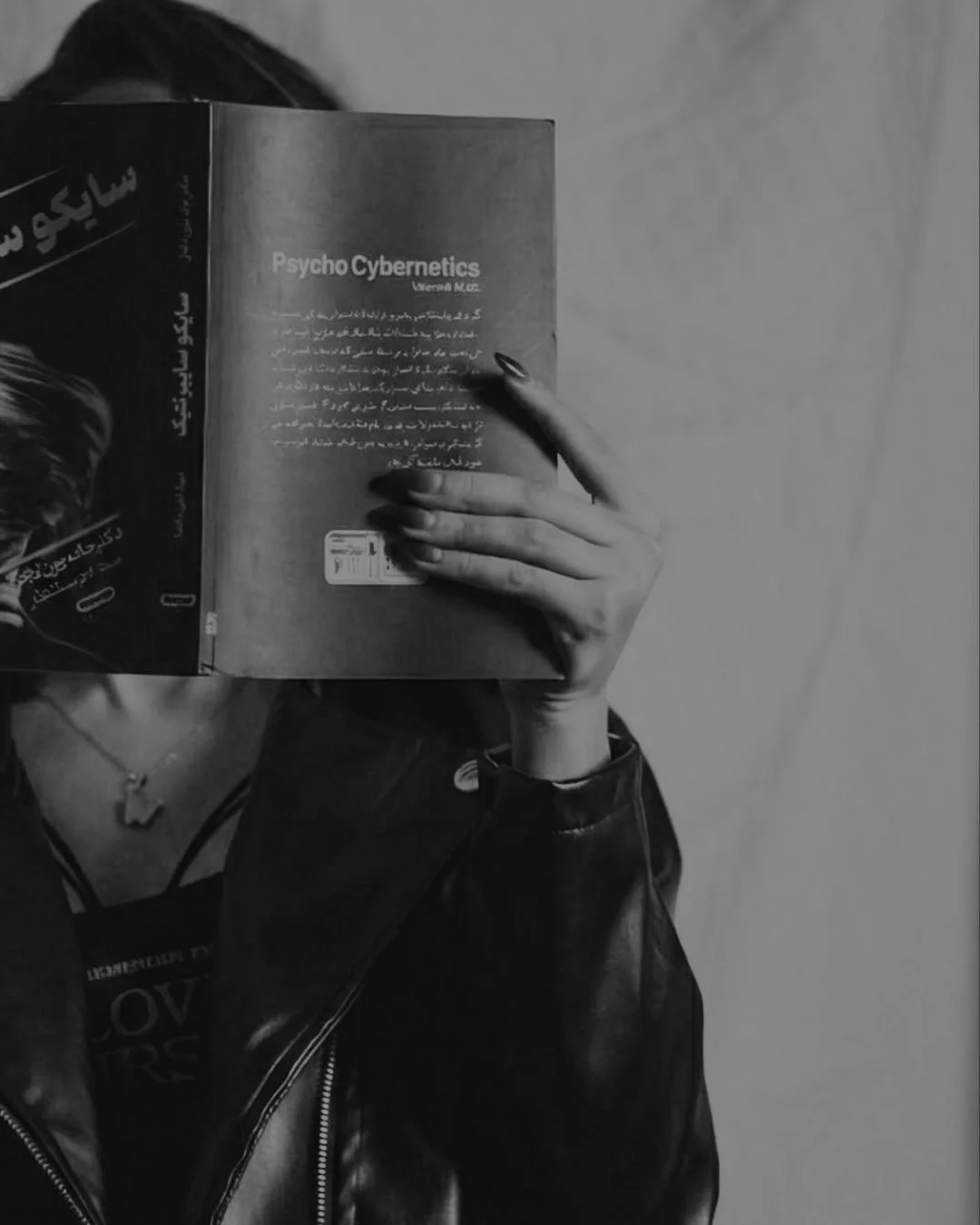 A person holding a book titled 'Psycho Cybernetics' covering their face, with a visible tattoo on their wrist and the person wearing a leather jacket, standing against a plain wall.