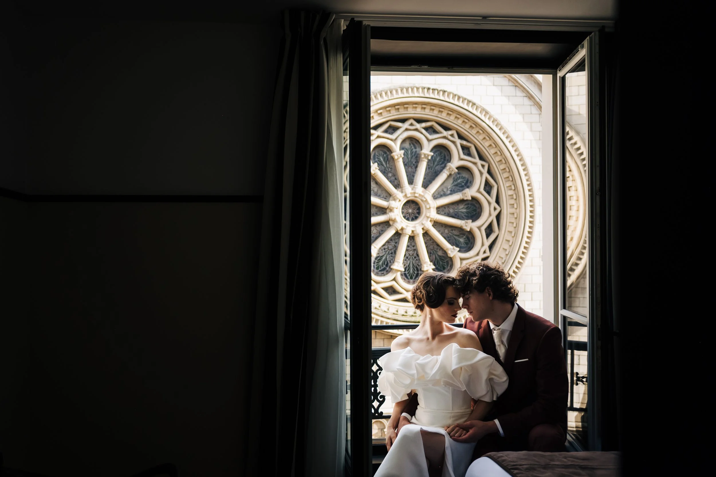A couple in wedding attire sitting on a bed near a window, with an ornate circular stained glass window in the background.