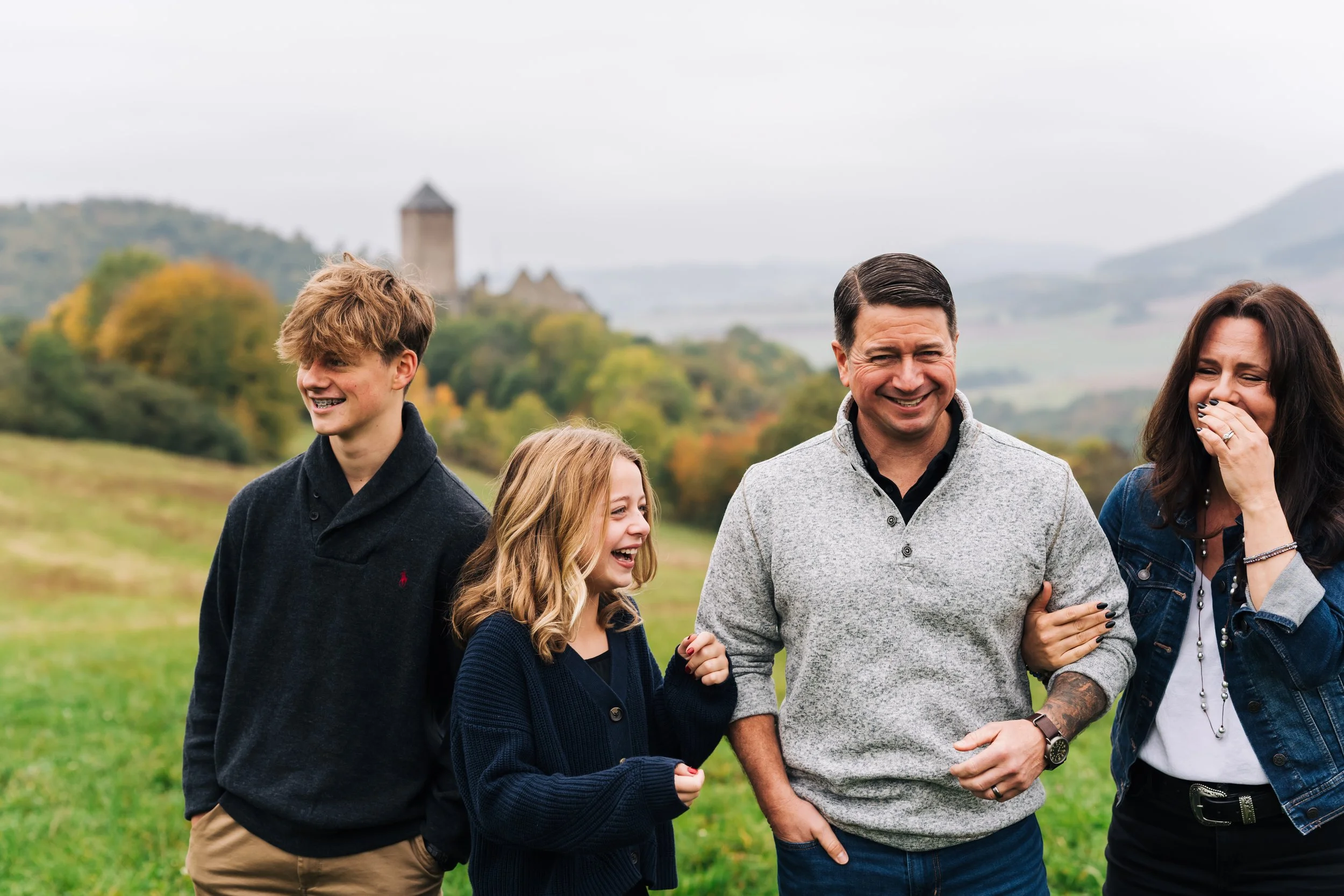 A family of four laughing outdoors in a green field with trees and a castle in the background.