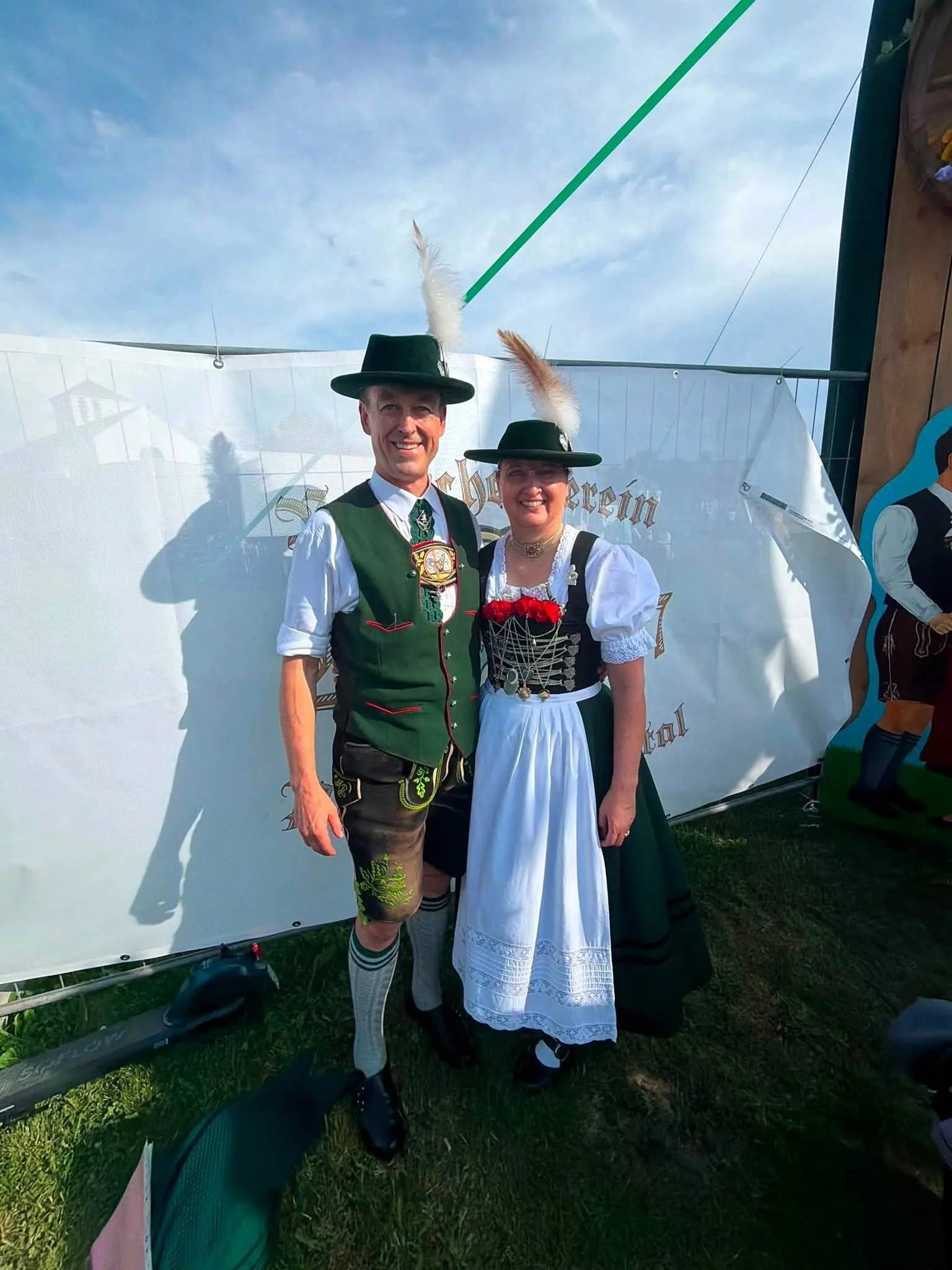 A man and a woman dressed in traditional German clothing, standing outdoors at an event, smiling. The man wears a green vest, lederhosen, and a hat with feathers, while the woman wears a dirndl dress with a lace apron and a hat with a feather.