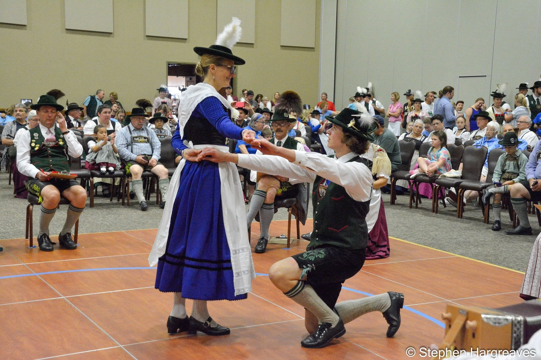 A woman dressed in traditional Bavarian clothing and a man kneeling on one knee in the final figure of a Schuhplattler dance. They are in a large indoor event space with many people wearing traditional Bavarian attire watching the dance.