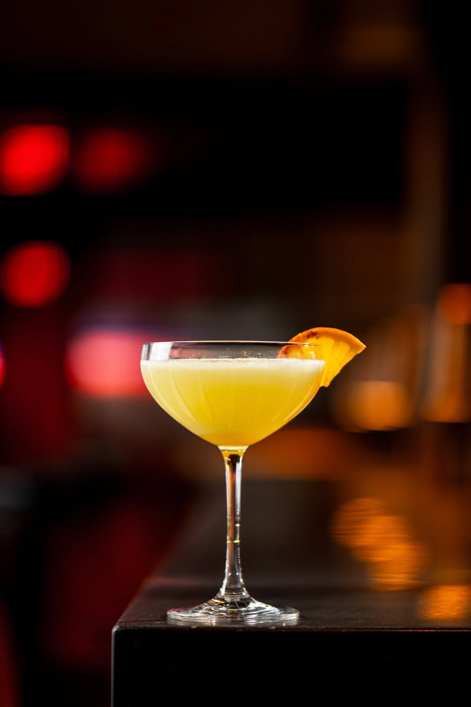 A yellow cocktail in a coupe glass garnished with an orange slice, set against a dark blurry background.