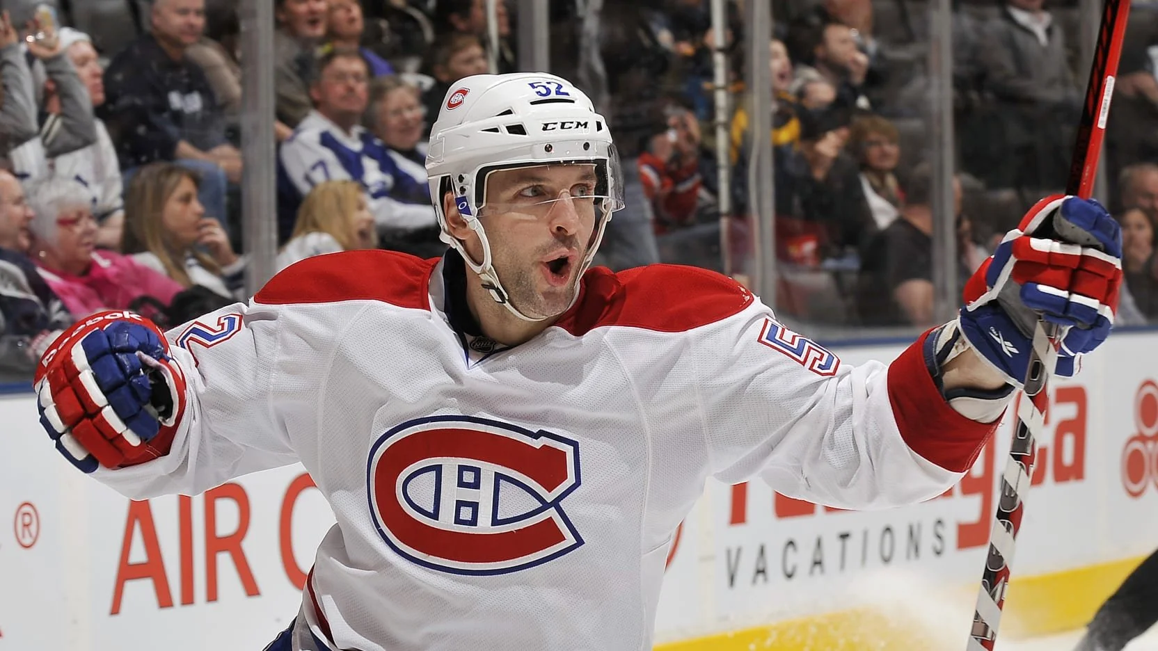 An ice hockey player wearing a Montreal Canadiens jersey celebrating on the ice rink with fans in the background.