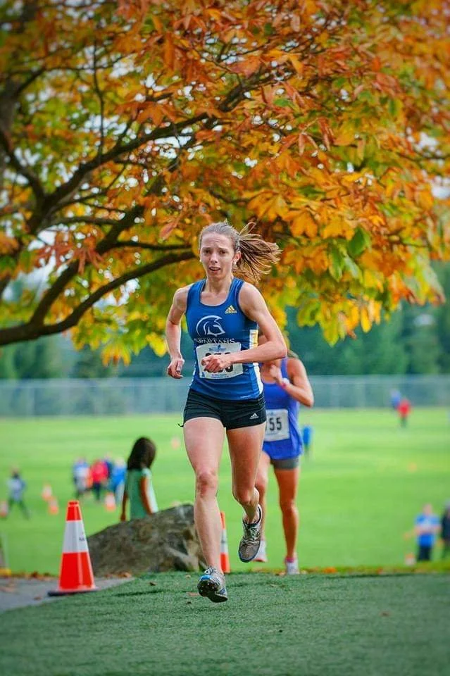 A young female athlete running in a race outdoors on a grassy field. She is wearing a blue athletic top and black shorts, with a participant number pinned to her top. The background features trees with autumn-colored leaves and other runners visible behind her.