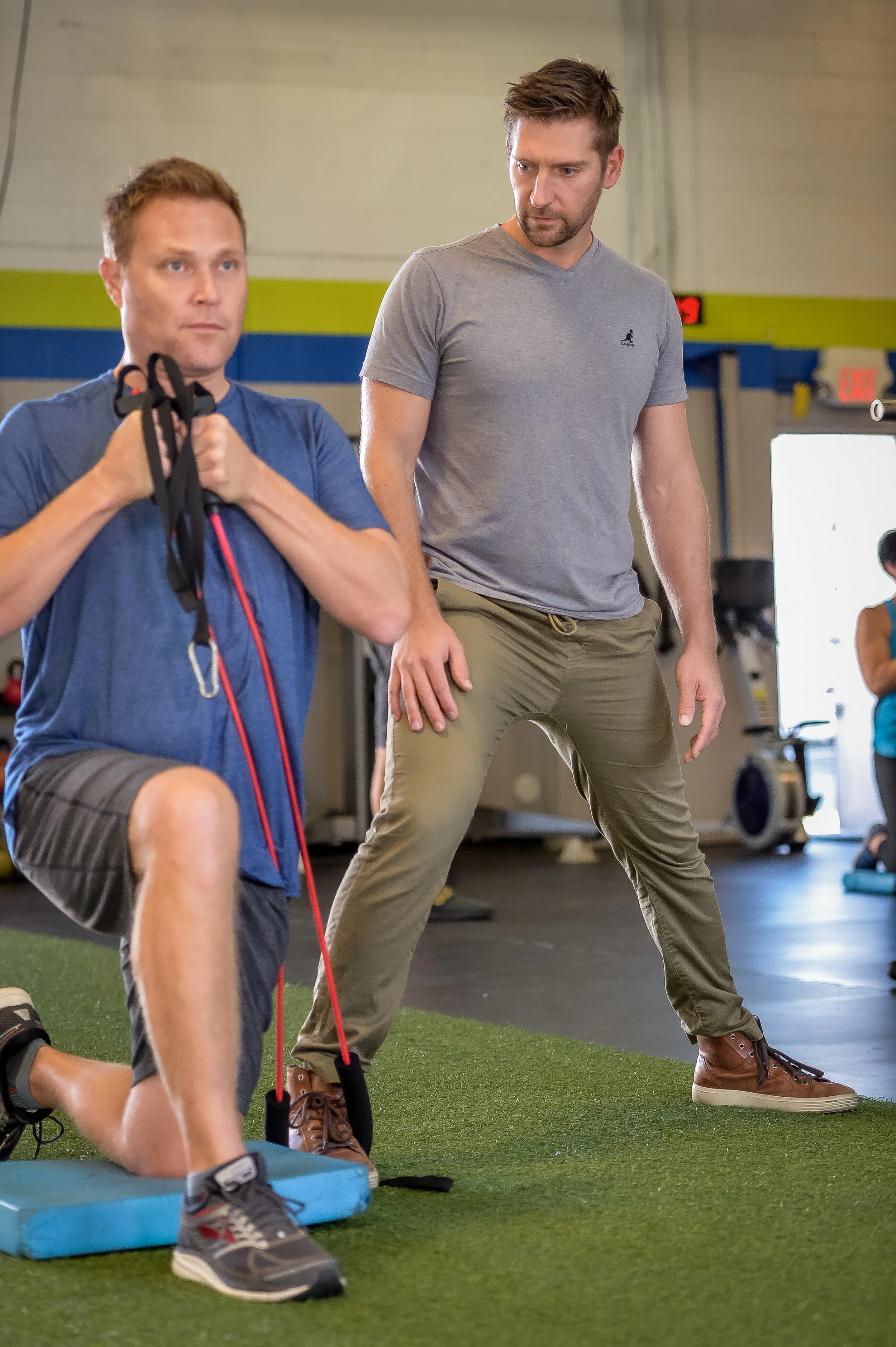 Two men in a gym performing lunges; one holding resistance bands, the other observing.
