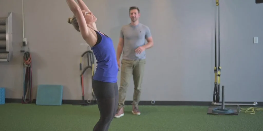 A woman stretching her arms above her head while a trainer observes in a gym.