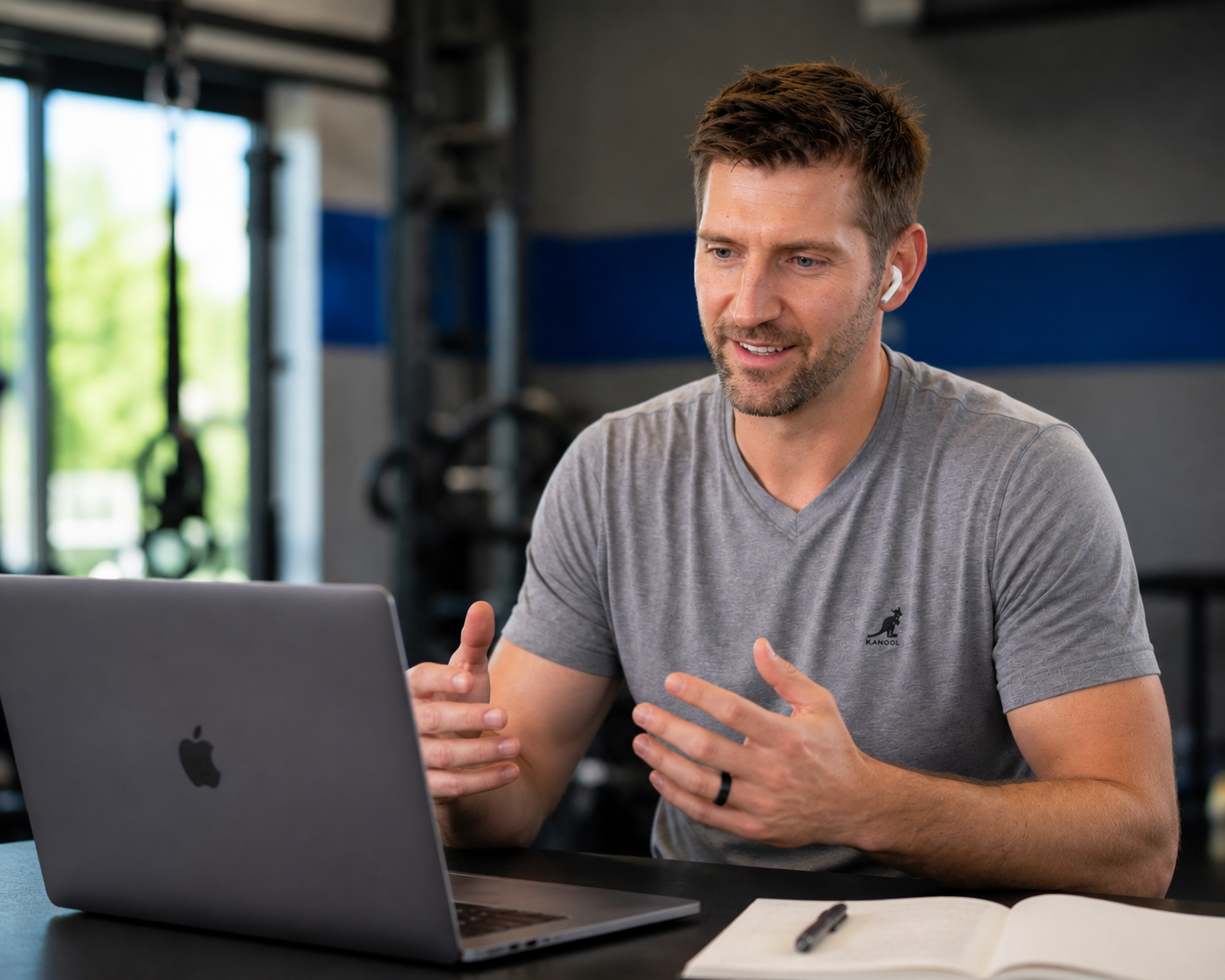 Darren McConaghy, sitting at a table with a silver MacBook in front of him, speaking during a video call in a gym or fitness center. Virtual Coaching