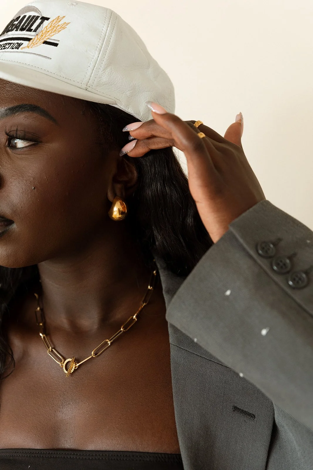 Close-up of a woman wearing a white hat with embroidery, gold jewelry including earrings and a necklace, and a gray blazer, touching her hat with her hand.