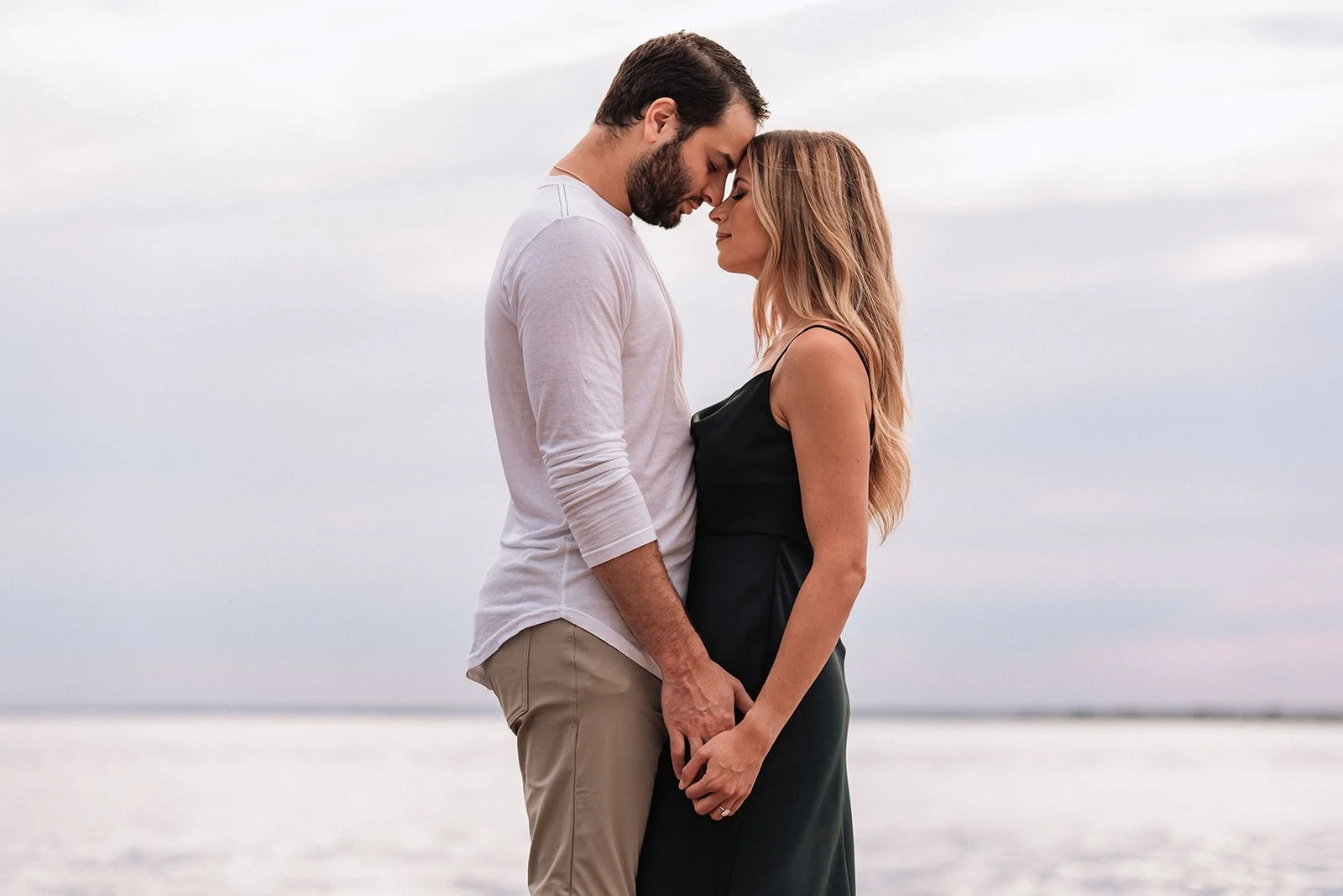 A couple stands close together on the beach, touching foreheads and holding hands. The man is wearing a white long sleeve shirt and khaki pants; the woman is in a black sleeveless dress. The sky is cloudy and the water is visible in the background.
