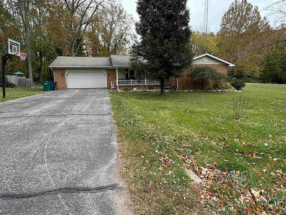 A house with a garage, a driveway, a front lawn with fallen leaves, and a basketball hoop to the left. There are trees in the background.