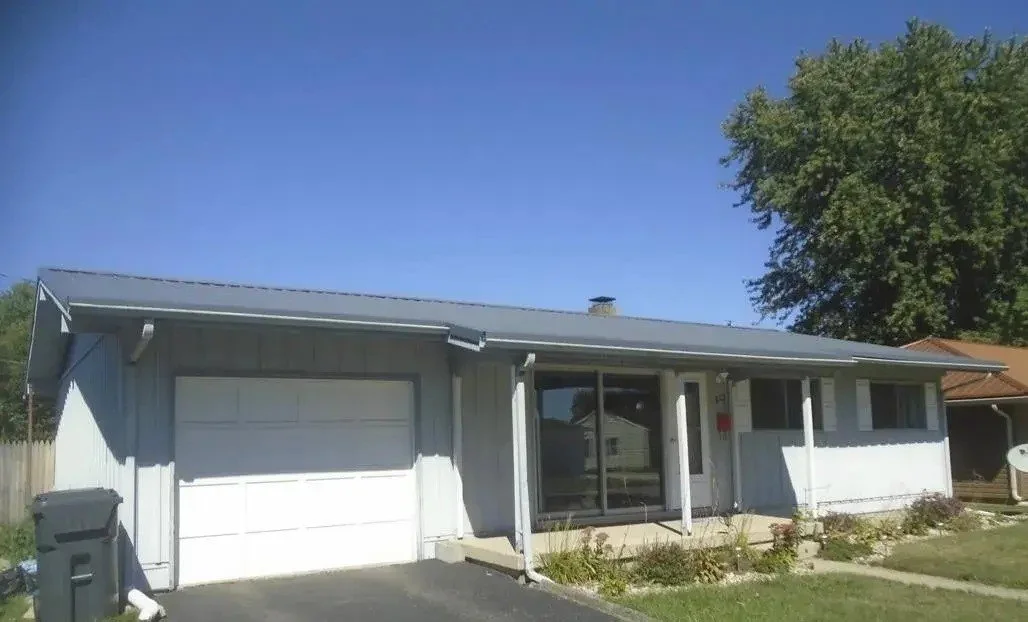 Single-story house with a white exterior, attached garage, sliding glass door entrance, small front yard with plants, and a large tree on the right, set under a clear blue sky.
