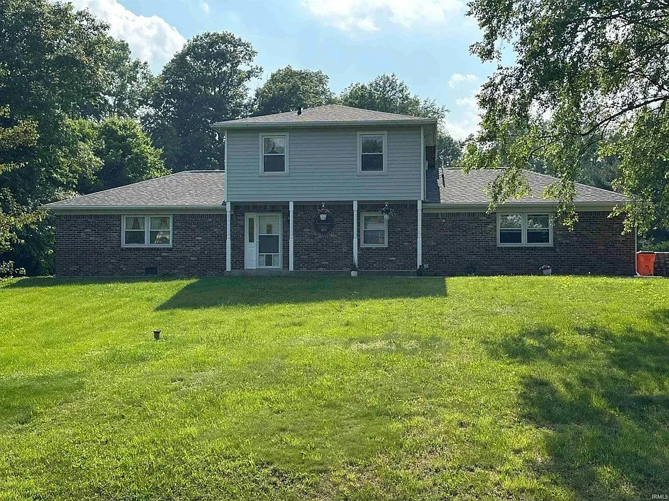 Two-story house with brick and siding exterior, open backyard, lush green lawn, trees in the background, and a partly cloudy sky.