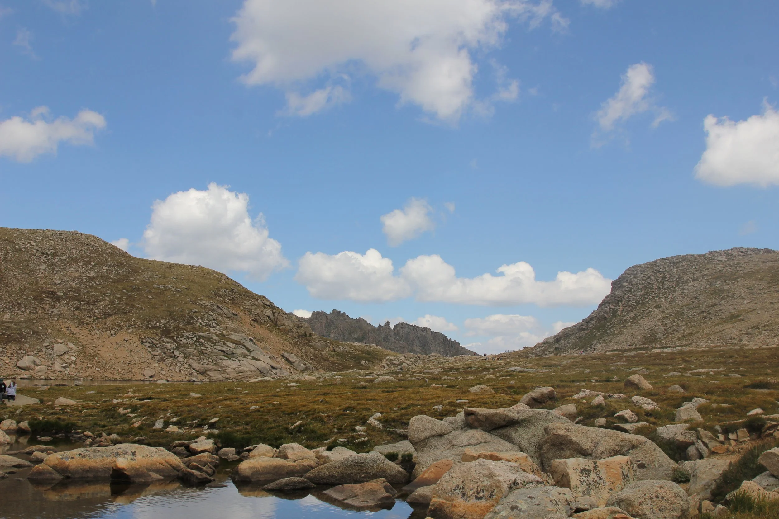 Scenic mountain landscape with rocky terrain, a small reflective water body, grassy plains, and a blue sky with scattered clouds.