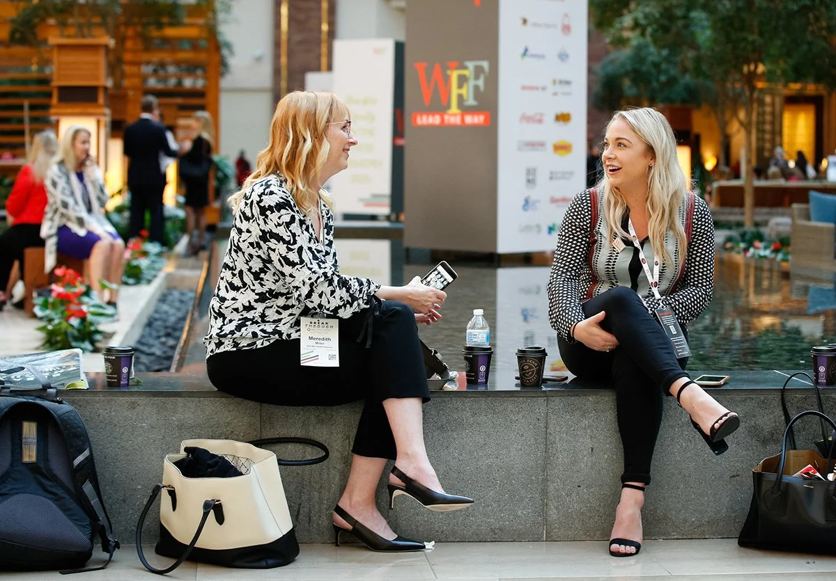 Two women with conference badges sitting and chatting by a water feature during a professional event, surrounded by bags and coffee cups.