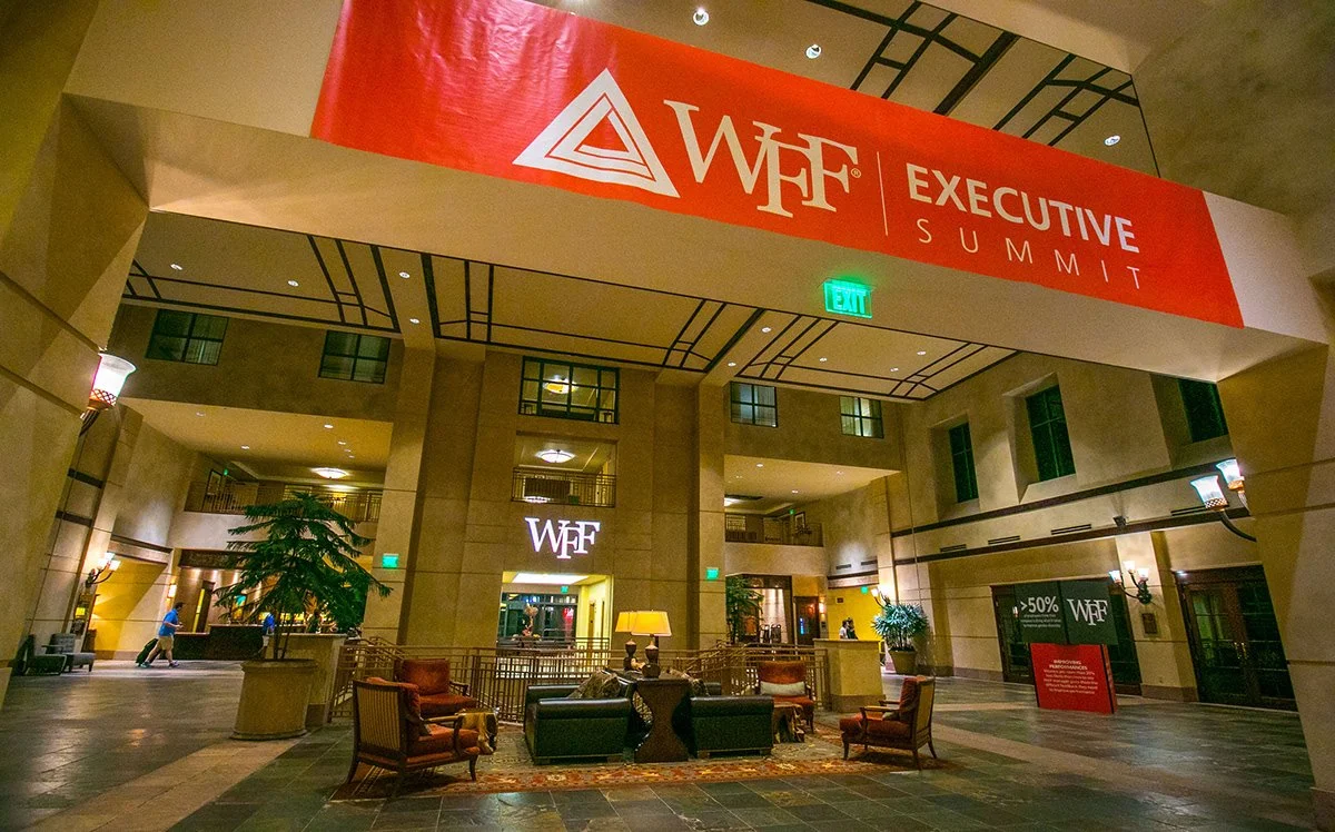 Hotel lobby with seating area, large plants, and a red banner hanging from the ceiling that says 'WFF Executive Summit.'