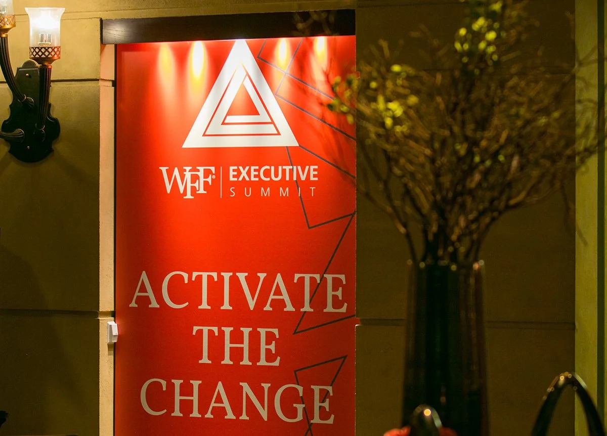 Red vertical banner with white text reading "Activate the Change" and logo for the WEF Executive Summit, with a geometric triangular symbol above the text, located on a wall near a lit wall sconce and a tall vase with branches.