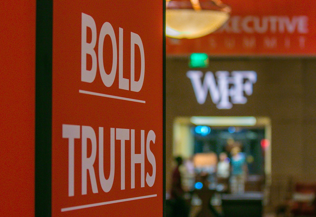 A sign that reads "BOLD TRUTHS" with a blurred background showing a restaurant or café with a sign that says "WFE".