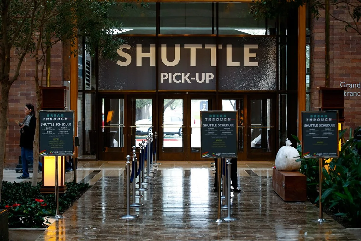 Indoor shuttle pick-up area with signs displaying shuttle schedule and route, and a person using a mobile device on the left side.