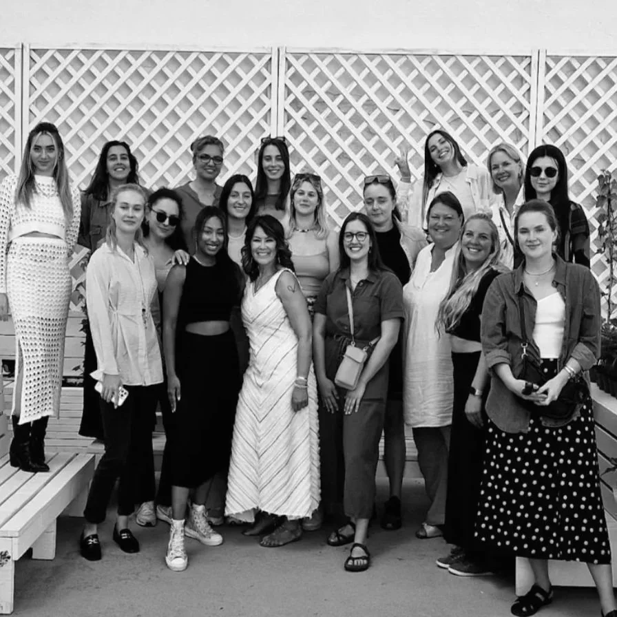 Black and white group photo of 15 Fortuna Network members with founder Daniella Jasmine against latticed wall
