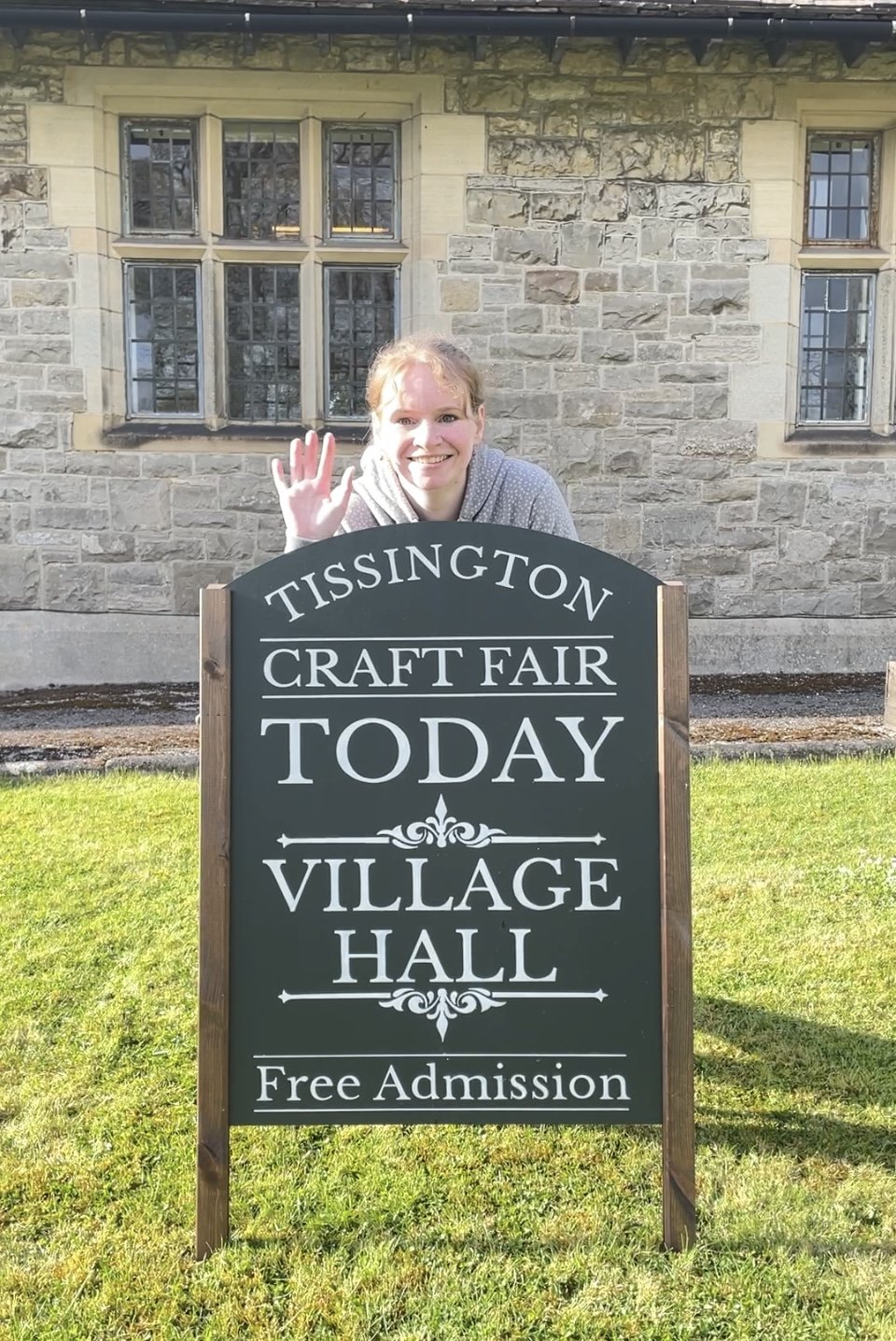 organiser of Tissington Craft Fairs standing behind their sign waving.