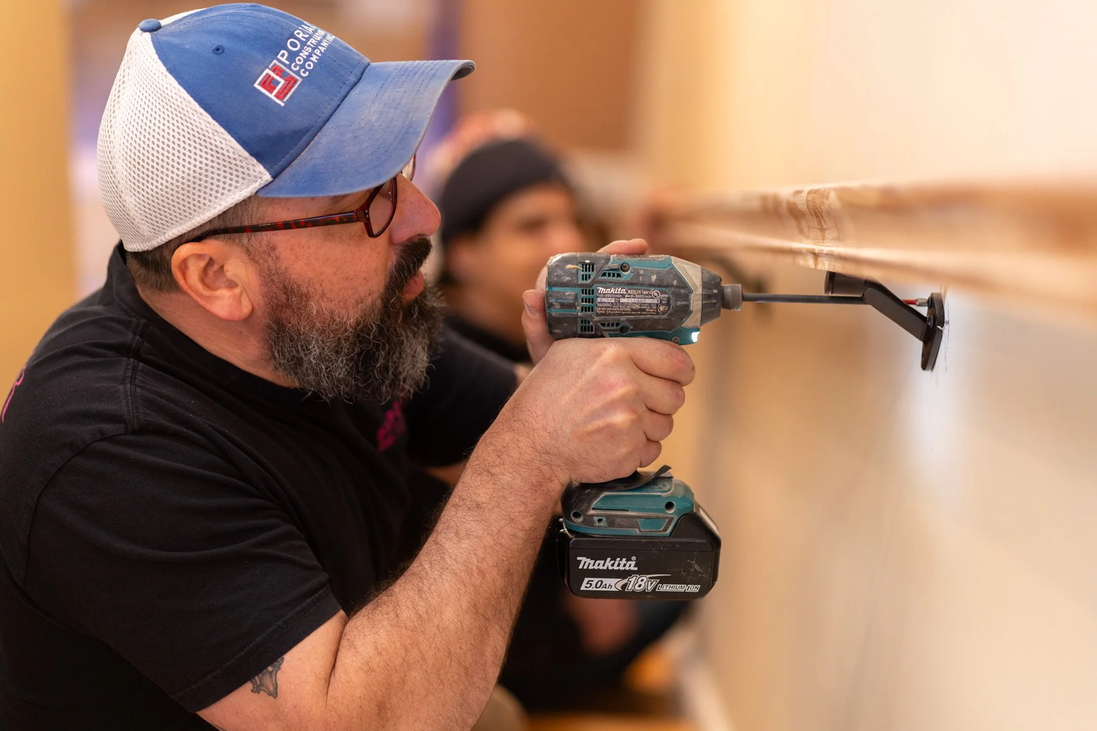 A man wearing glasses and a blue cap with white mesh is using a cordless drill on a wooden surface. There are blurred people in the background.