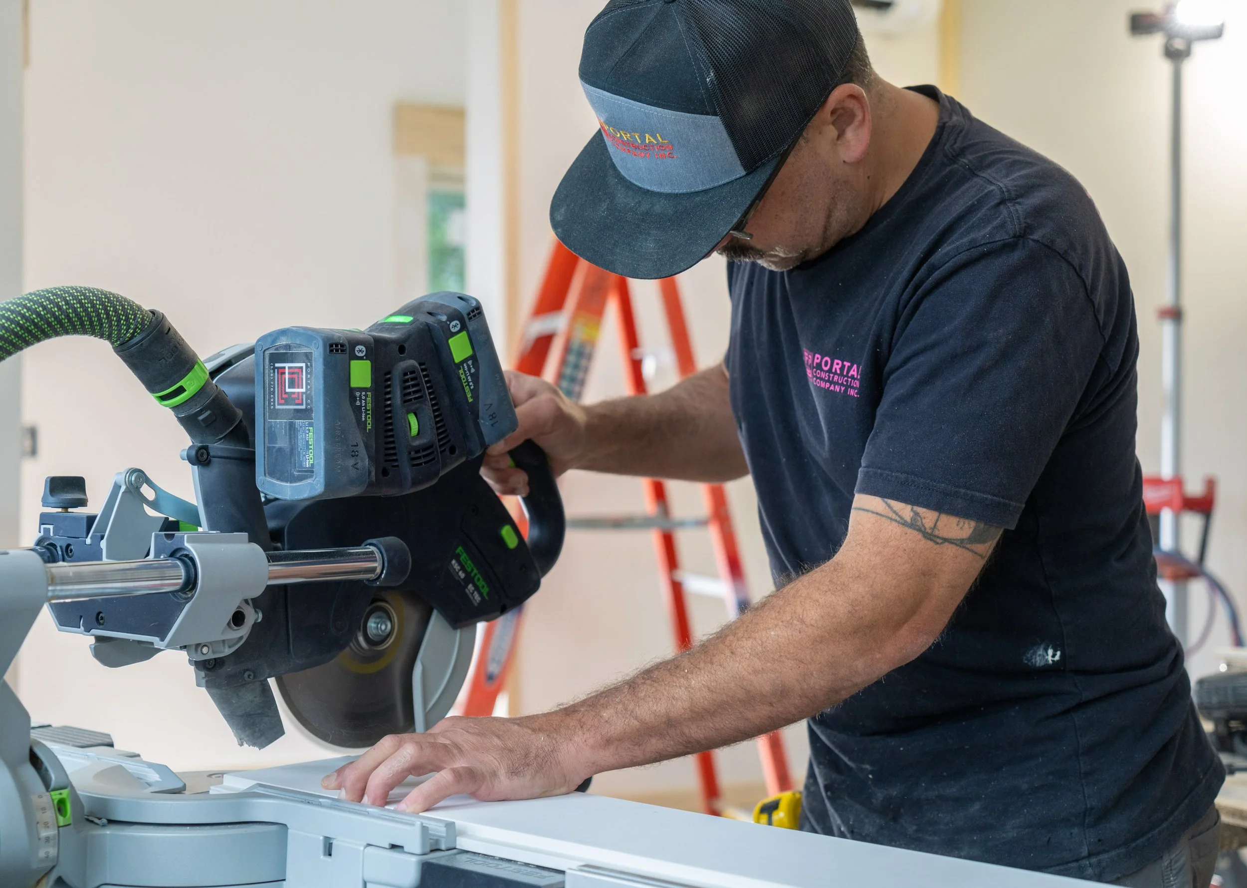 A man wearing a black cap and t-shirt is working with a large chop saw in a construction setting. There are ladders and construction equipment in the background.
