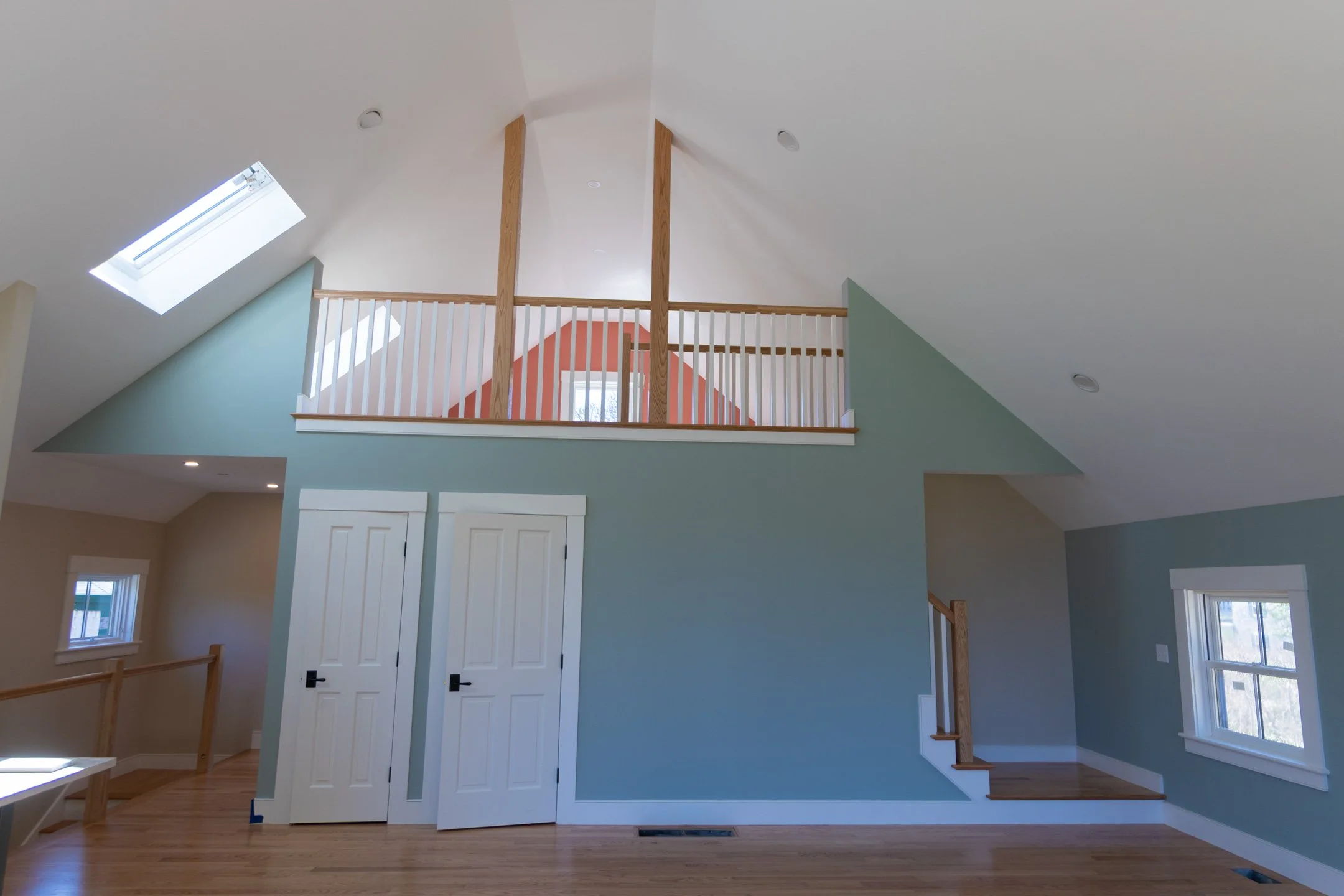 Interior view of a spacious room with a high vaulted ceiling, skylights, a staircase with a wooden railing, and hardwood flooring painted walls in shades of light green and peach.