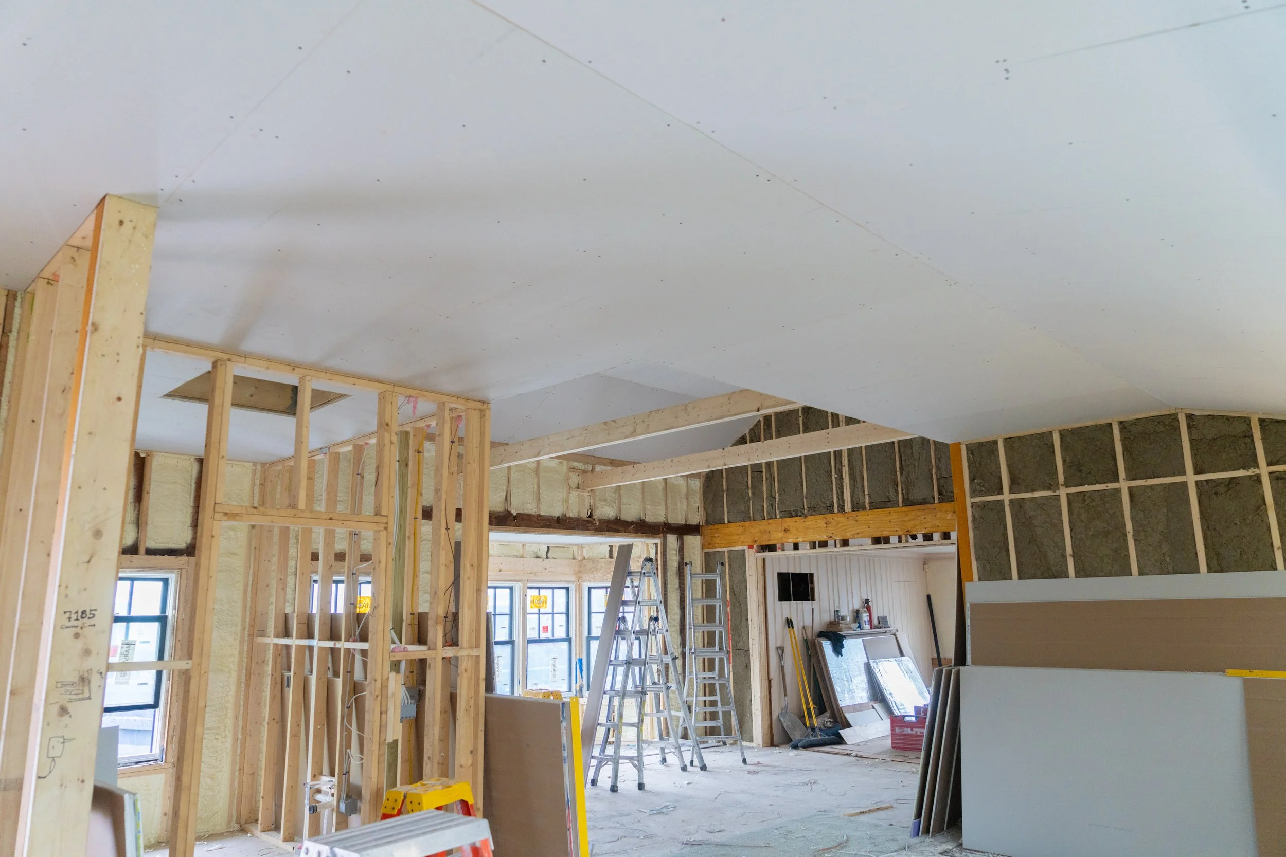 Interior view of a house under construction with wooden framing, drywall, ladders, and building materials visible.