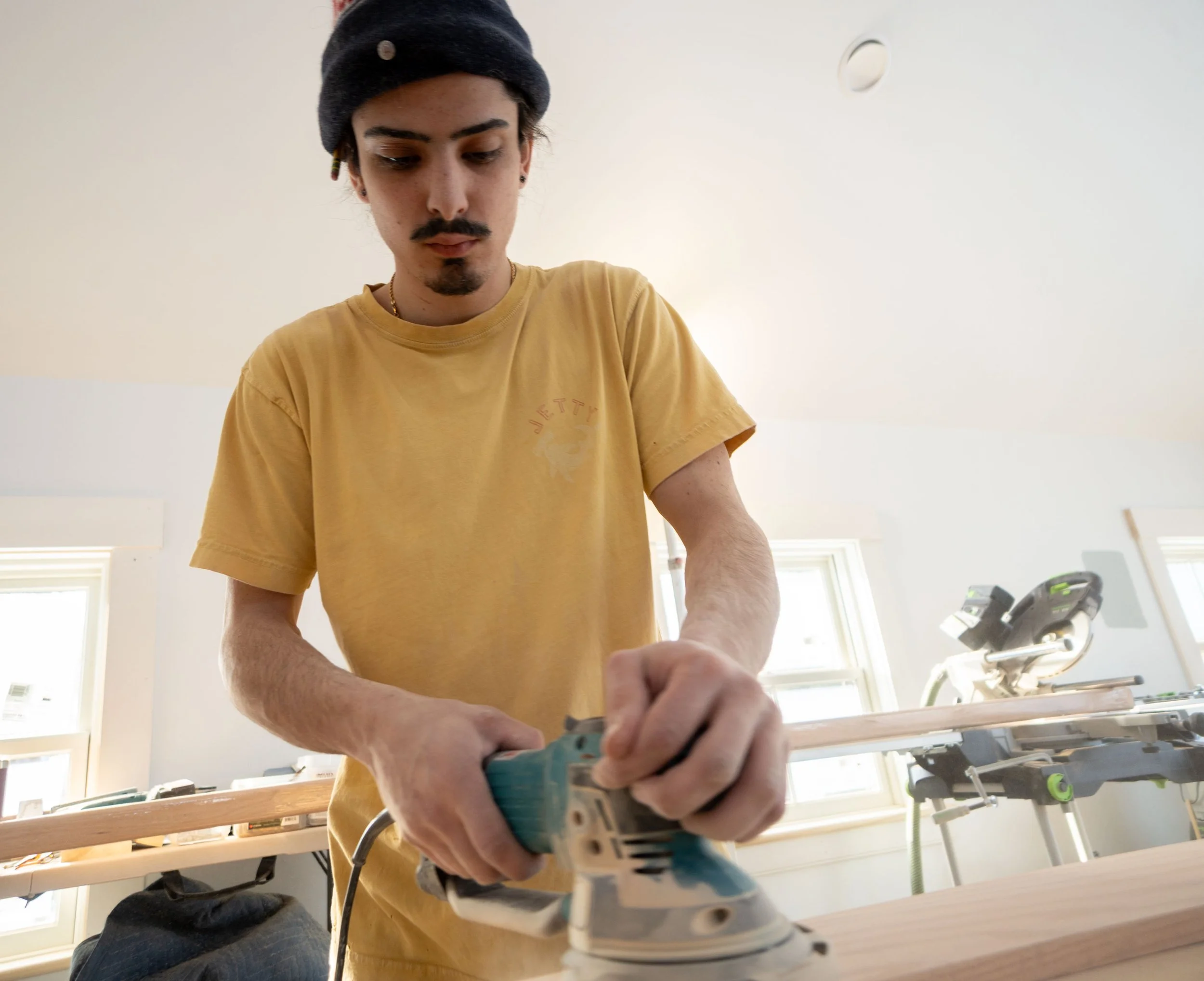 Young man with dark hair, mustache, and earrings operating a power saw while working on a piece of wood in a well-lit room with large windows.