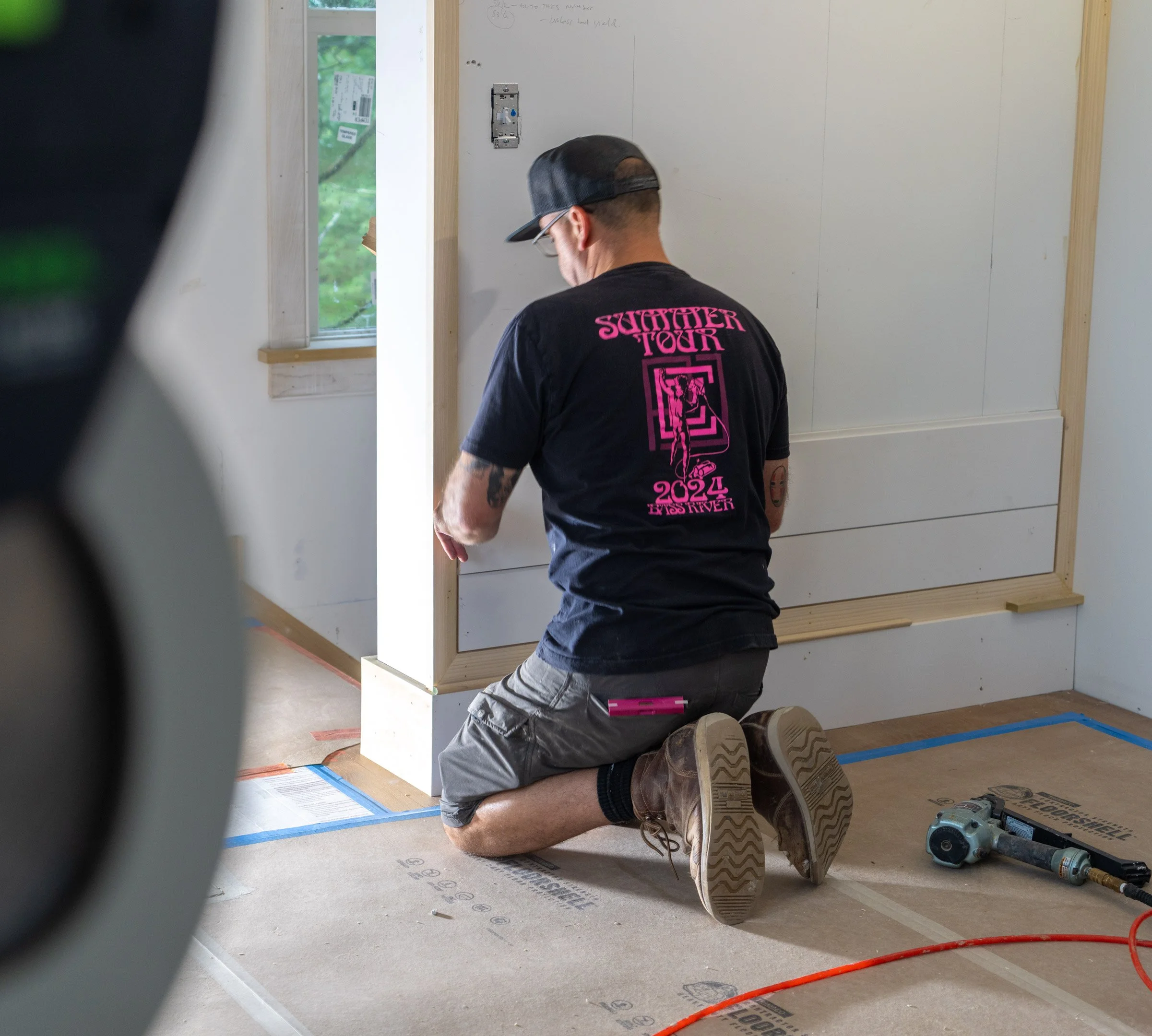 A person kneeling on the floor of a room under construction, working on an electrical outlet or switch on the wall. The person is wearing a black t-shirt with pink writing and graphics, shorts, and work boots. Various tools and construction materials are scattered on the floor around them, and a window shows green trees outside.